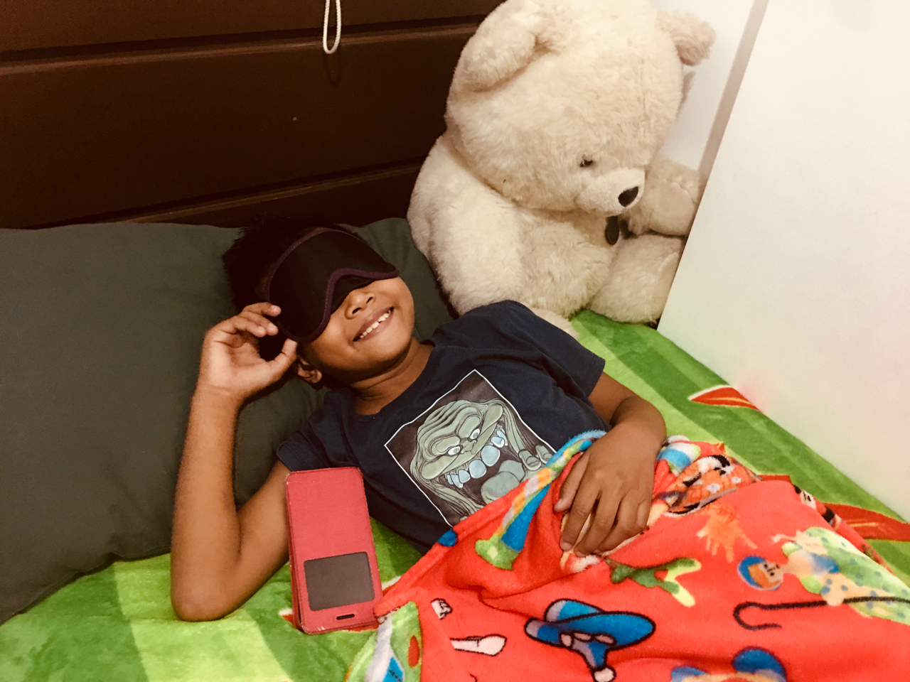 a boy wearing bat mask in bed with teddy bear